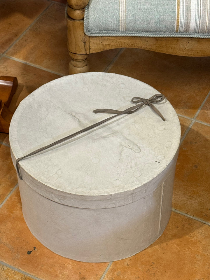 Cappelliera vintage cilindrica in cartone rivestito grigio chiaro con coperchio removibile e chiusura a nastro.