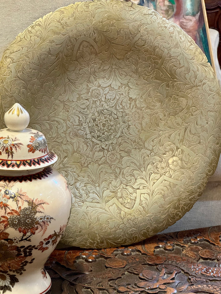 Grande centrotavola circolare in metallo dorato con elaborato decoro floreale e piedini inferiori.