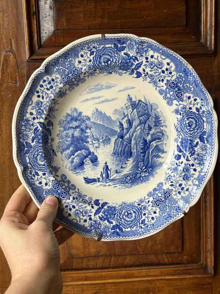Piatto da parete in ceramica del noto marchio Villeroy &amp; Boch decoro "Burgenland".

Il piatto presenta una scena centrale paesaggistica che raffigura un tranquillo paesaggio fluviale con un castello arroccato su una collina, circondato da alberi e montagne sullo sfondo. Un'imbarcazione sul fiume e figure umane in primo piano contribuiscono a creare un'atmosfera bucolica e serena, tipica del romanticismo paesaggistico. Il tutto è circondato da un elaborato motivo floreale.
Misure 25 cm