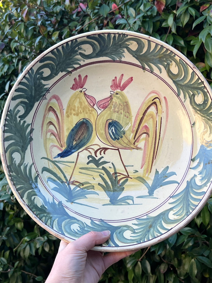 Piatto in ceramica decorato a mano realizzato dall'artista Carlo Zubrod Grottaglie. Il decoro raffigura una scena vivace e colorata con galli stilizzati, circondata da un decoro fogliaceo ornamentale. Lo stile pittorico richiama la tradizione artigianale, valorizzando ogni dettaglio con autenticità.

Diametro 33 cm