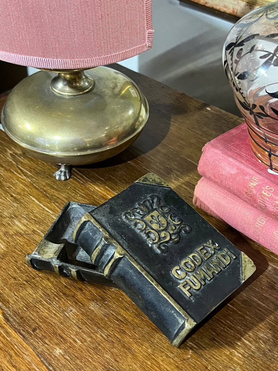 Originale Posacenere vintage realizzato in bronzo a forma di libro antico. Il design include dettagli in rilievo come uno stemma e l'iscrizione "Codex Fumandi", che aggiungono un tocco di fascino storico al pezzo.

In entrambi i lati sono presenti due poggiasigarette.

Misure 7x9x3 cm