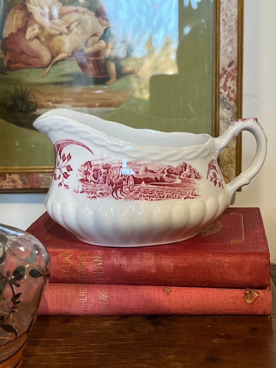Deliziosa salsiera in porcellana realizzata dalla rinomata tradizione Staffordshire, Inghilterra. Il pezzo è decorato con un'affascinante scena rurale in tonalità rosso rubino, rappresentante il paesaggio di "Hampstead Heath", un classico esempio dello stile inglese tradizionale.

Un'elegante aggiunta per una tavola in stile vintage o per gli amanti dell'antiquariato inglese.

Misure 17x11x10cm&nbsp;