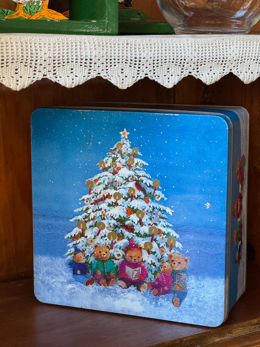 Scatola di latta natalizia vintage blu con cinque orsetti seduti nella neve davanti a un albero di Natale illuminato, dimensioni 23 × 23 cm.