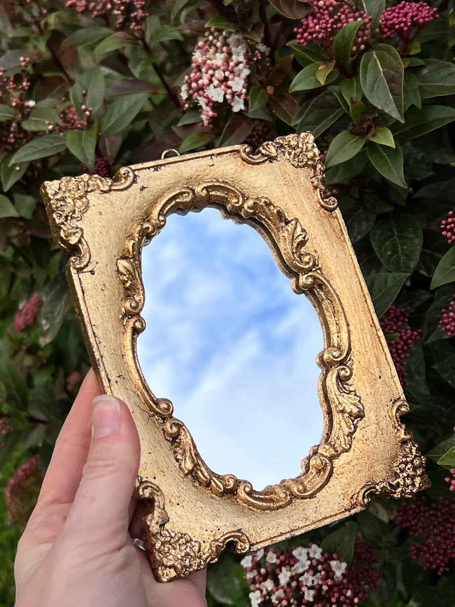 Specchietto di misure 16x12 cm, di ispirazione barocca con cornice intagliata e dorata a foglia oro. Lunga tutta la cornice è presente un delizioso decoro con angoli floreali. Con una maestria che trascende il tempo, i nostri specchietti sono capolavori realizzati a mano che catturano l'essenza dell'eleganza barocca. Ogni cornice, finemente scolpita e rifinita con foglia d'oro, è frutto di tecniche di antichizzazione che regalano un'anima vissuta e senza tempo ad ogni pezzo.