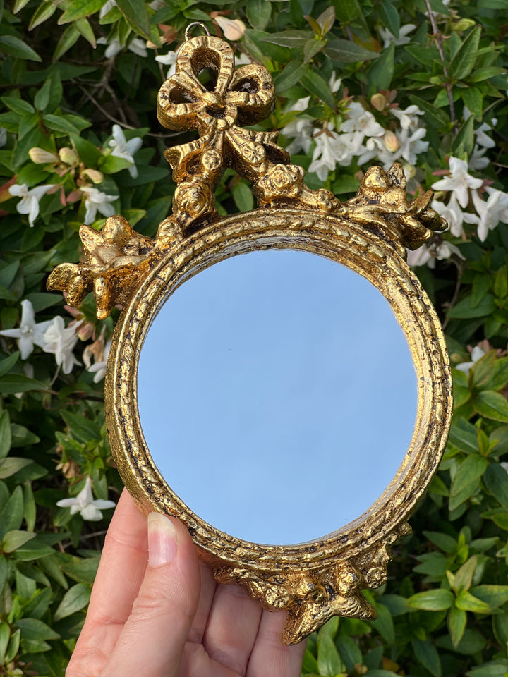 Specchietto di misure 13x19 cm, di ispirazione barocca con cornice intagliata e dorata a foglia oro. Nella parte superiore è presente un elaborato motivo a fiocchetto.

Con una maestria che trascende il tempo, i nostri specchietti sono capolavori realizzati a mano che catturano l'essenza dell'eleganza barocca. Ogni cornice, finemente scolpita e rifinita con foglia d'oro, è frutto di tecniche di antichizzazione che regalano un'anima vissuta e senza tempo ad ogni pezzo.