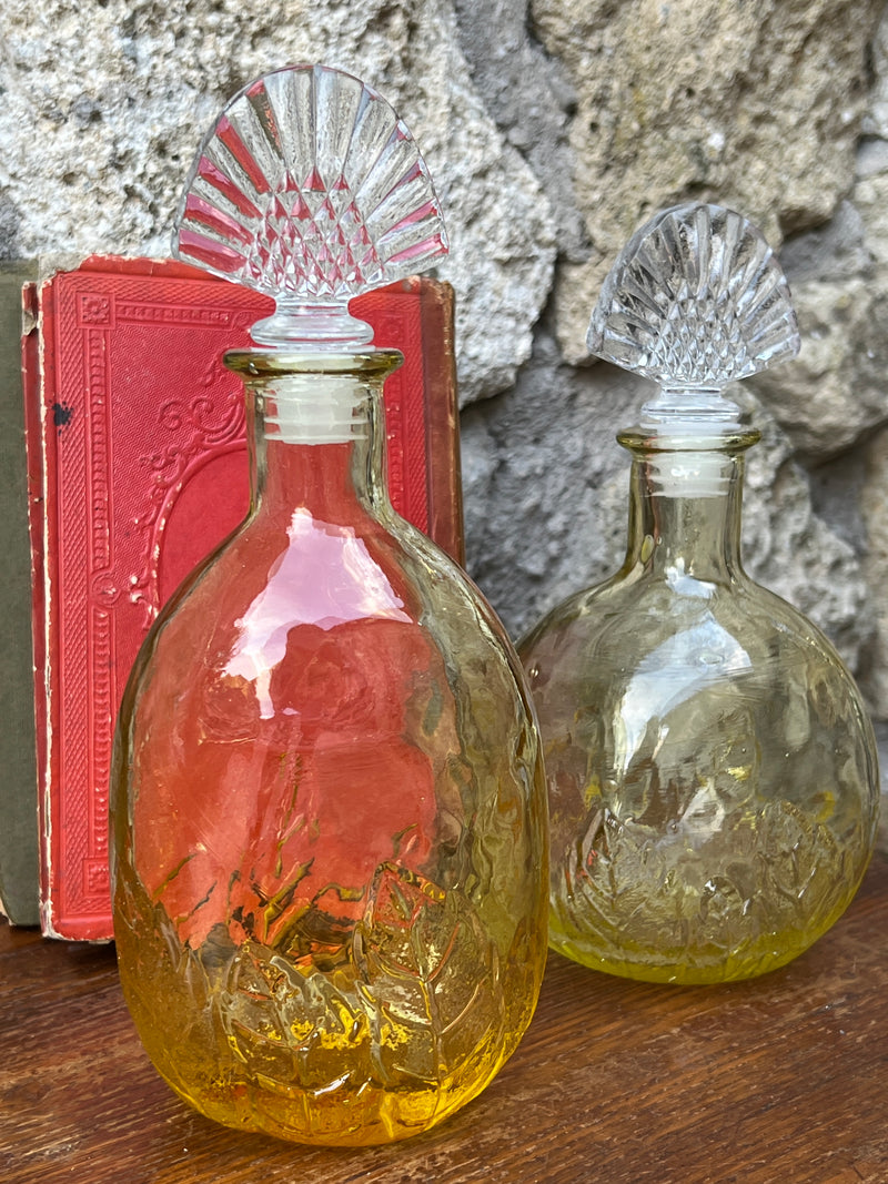 Bottiglia in vetro di colore giallo con tappo in vetro trasparente, di forma ovale.  Presenta una decorazione floreale sulla parte inferiore della bottiglia.