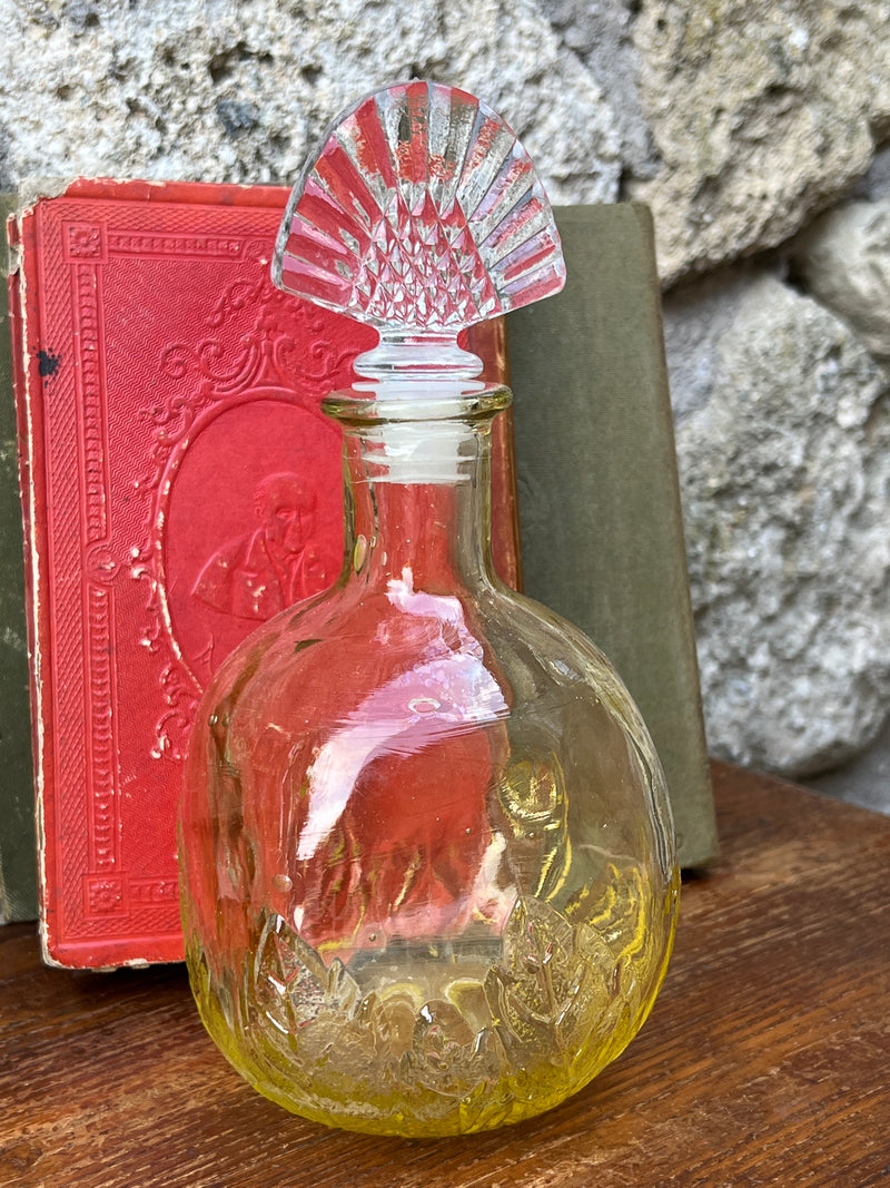 Bottiglia in vetro di colore giallo con tappo in vetro trasparente, di forma squadrata con gli angoli smussati.  Presenta una decorazione floreale sulla parte inferiore della bottiglia.
